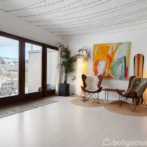 Stue med to stole foran et farverigt abstrakt maleri, omgivet af planter. Store glasdøre åbner til en snedækket terrasse. Loftet er dekoreret med lyskæder.