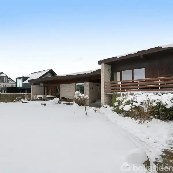 Et moderne hus med træfacade ligger stille i et snedækket landskab. Omgivet af andre boliger i en rolig boligområde.