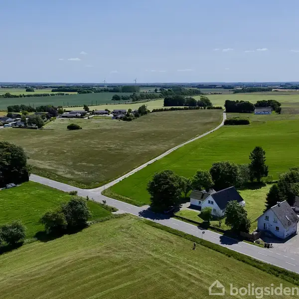 Landsbyhuse står stille ved en vej, omgivet af grønne marker og træer under en klar blå himmel. I baggrunden ses flere spredte huse og åbne marker.