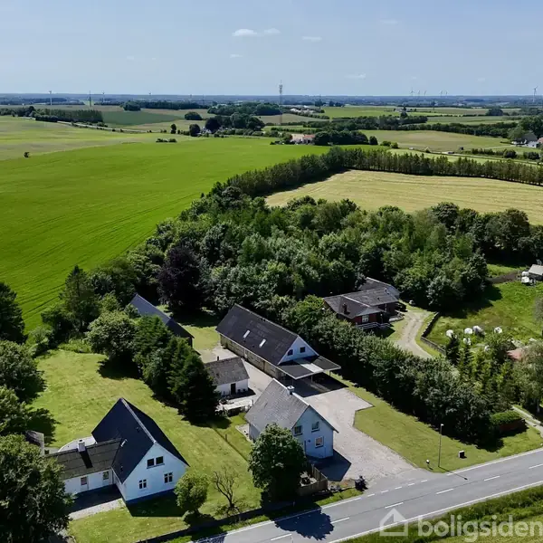 Et område med huse ligger langs en vej omgivet af grønne marker og træer. Bebyggelsen er placeret i et åbent, landligt landskab med klare himmel og frit udsyn.