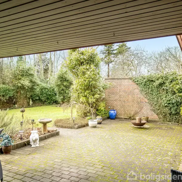 Overdækket terrasse i have, omringet af murstensvægge og buske. Terrassen er dekoreret med potteplanter, havemøbler og en fuglebad i midten. Græsplæne ses i baggrunden.