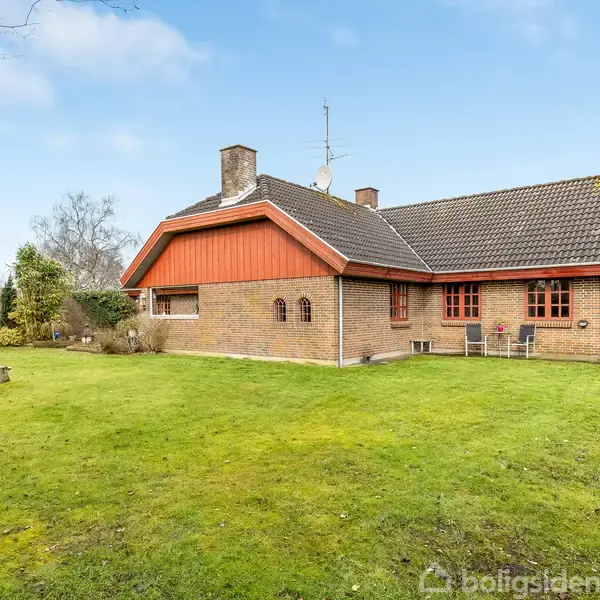 Et murstenshus med rødt tag står på en velholdt græsplæne. Huset har flere vinduer og en rød dør. Haven er omkranset af træer og buske under en klar, blå himmel.