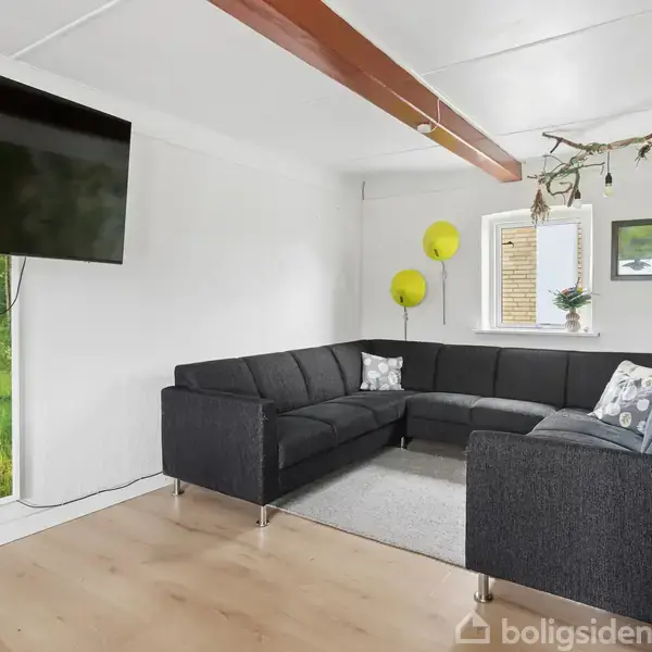 Sort hjørnesofa står i stuen, omgivet af hvidmalede vægge og et træagtigt loftselement. Et stort fladskærms-TV hænger på væggen, og en lampe og grønne dekorationer skaber en hyggelig atmosfære.