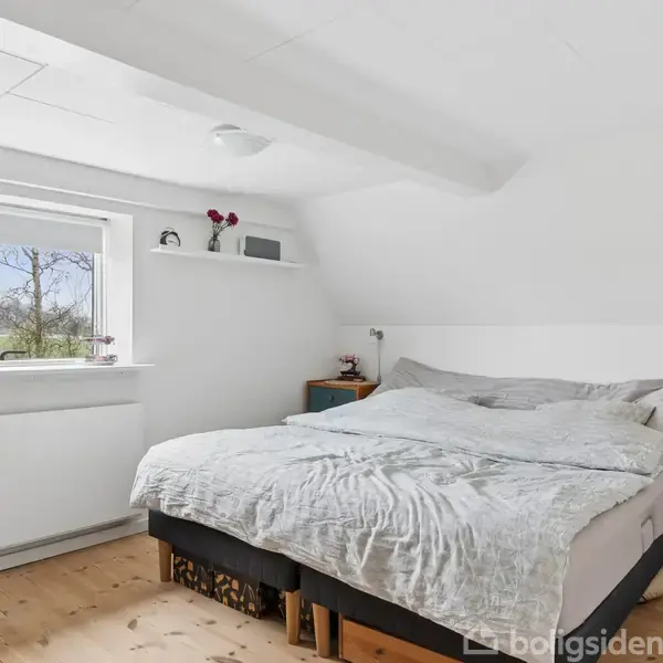 Seng med gråt sengetøj står i et lyst soveværelse. Der er en kommode ved siden af sengen og et vindue med udsigt til træer udenfor. En radiator er under vinduet.