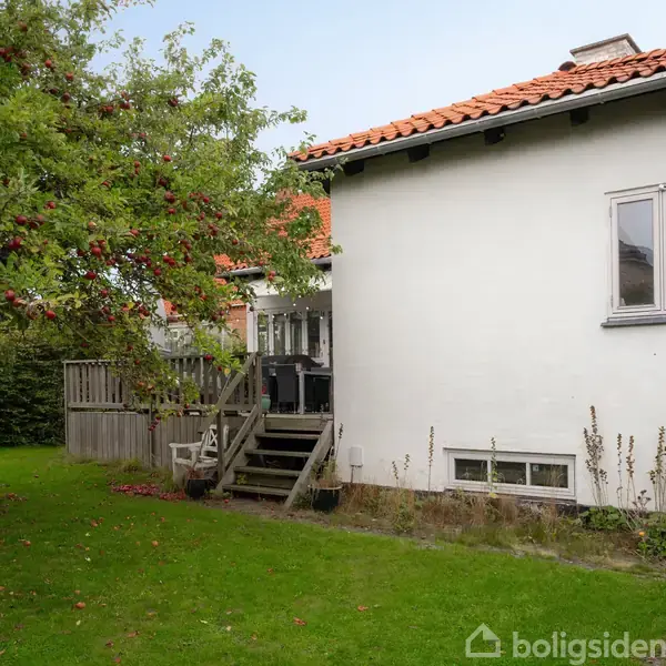 Hvidt hus med rødt tag står i en have med et æbletræ. Trappen fører til terrasse med gelænder. Græs og buske omgiver bygningen.