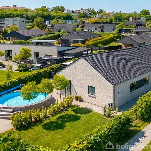 Et moderne enfamiliehus med gråt tag står i en velplejet have. En rund swimmingpool er placeret ved siden af huset. Baggrunden viser en række nabolagshuse omgivet af grønne områder.