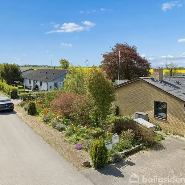 Et murstenshus omgivet af en have ligger ved en rolig vej. To nabohuse kan ses, samt en parkeret bil på vejen. I baggrunden findes marker med rapsblomster under en blå himmel.