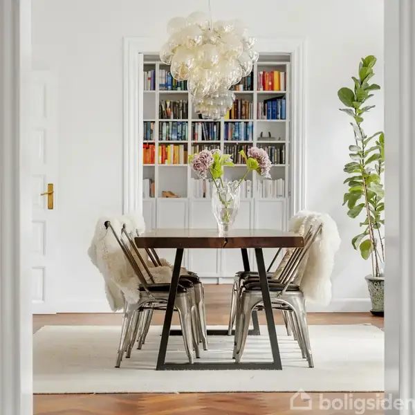 En spisebord med seks stole står i midten af et lyst rum, omgivet af en stor bogreol og en potteplante. Loftlampen hænger over bordet, som har en vase med blomster.