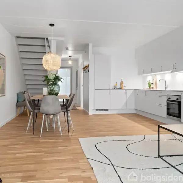 En moderne spisestue med et træbord og fire stole, en stor lampe over bordet, trætrappe i baggrunden, og et køkken til højre med hvide skabe og indbyggede hvidevarer.
