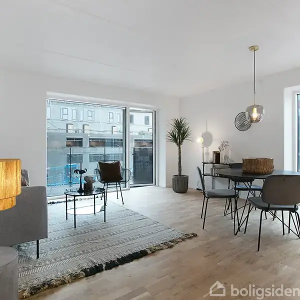 Stue med spisebord og stole står i midten. Store vinduer lukker lys ind. En plante og et gulvtæppe tilføjer hygge. Bygninger ses udenfor. Lampen hænger over bordet.