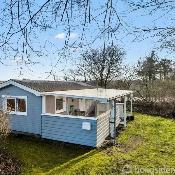 Et lille lyseblåt træhus står på en grøn græsplæne omgivet af træer. Huset har en overdækket terrasse og vinduer, der giver udsyn til en klar blå himmel.