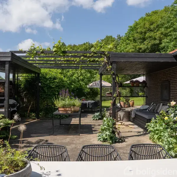 En terrasse har en pergola med klatreplanter og møbler, omgivet af blomsterbede og grønne planter. Et bord med stole står i forgrunden. Solrigt vejr skaber en hyggelig atmosfære.