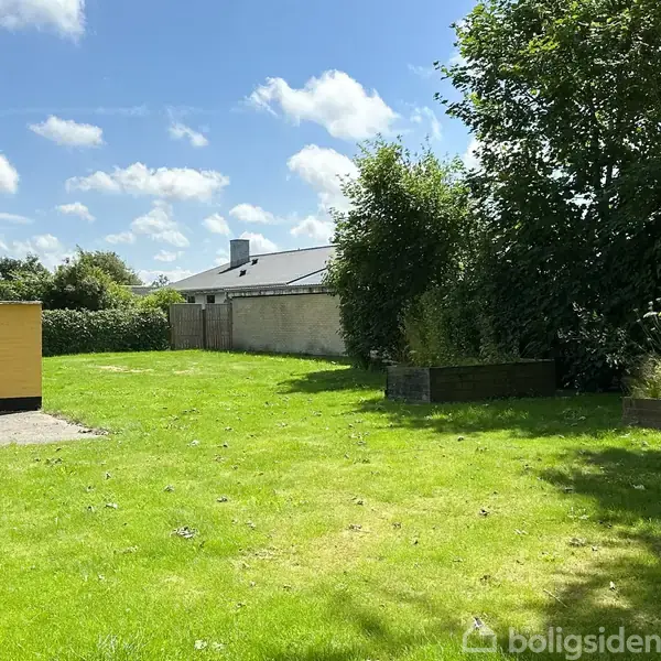 Græsplæne med skur til venstre og en bænk, omgivet af træer og buske, under en klar blå himmel med skyer.