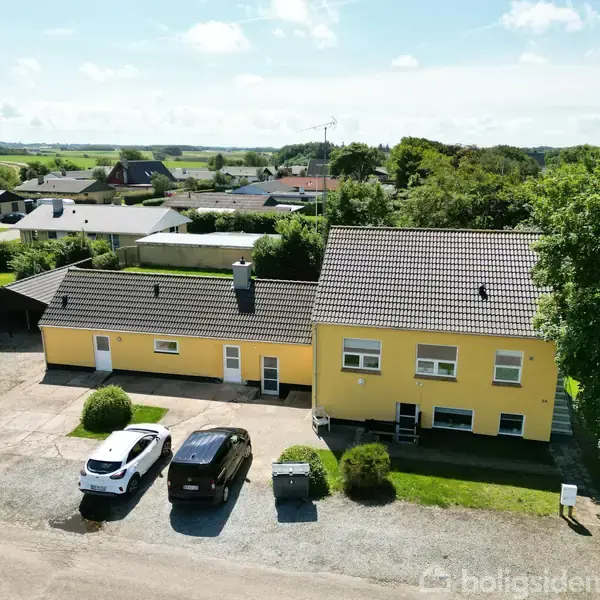 Gul bygning med to sektioner og gråt tag står på en grusbelagt gårdsplads. To biler er parkeret foran. I baggrunden ses en grøn have og træer samt flere huse.