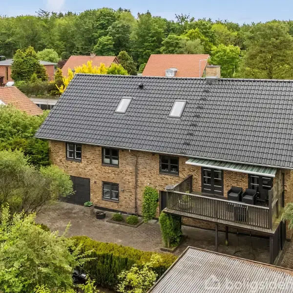 Et stort, rødt murstenshus med en grå tagterrasse udstyret med møbler. Det omkranses af frodige træer og buske i et grønt boligområde med flere huse i baggrunden.
