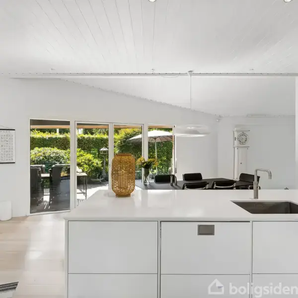 En moderne, hvid køkkenø med indbygget vask står i et lyst køkkenalrum. Gennem store vinduer ses en udendørs terrasse og grønne omgivelser. En kalender hænger på væggen.