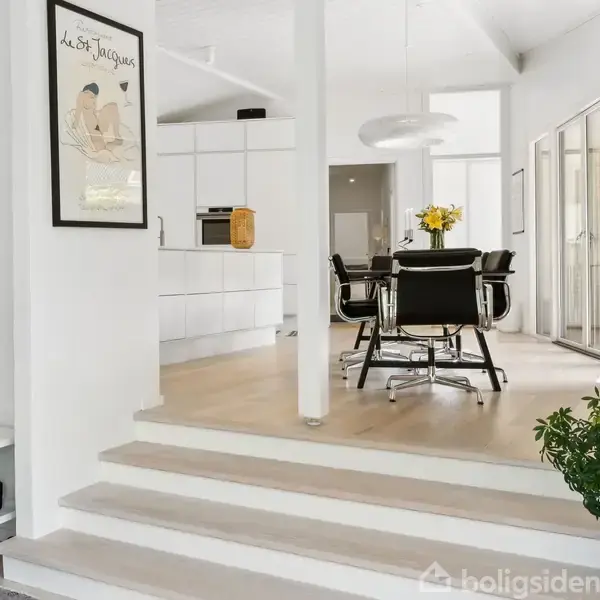 En moderne spisestue med et rundt bord og sorte stole er omgivet af hvide vægge og store vinduer. En plakat hænger på væggen med teksten "La Soirée Jacobs."