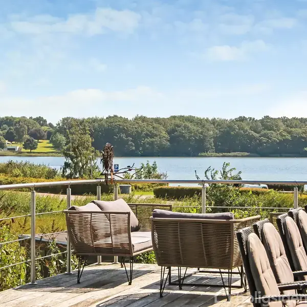 Terrasse med stole og borde vender mod en sø og skov i baggrunden under en klar himmel. Sollys skaber en hyggelig atmosfære.
