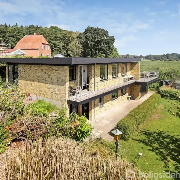 Toetagers moderne hus belagt med gule mursten, står blandt frodig natur. Husets store vinduer og terrasser vender mod grønne omgivelser, der omfatter en velplejet græsplæne og tætte buske.