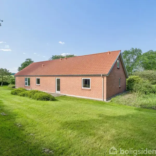 Et rødt murstenshus med mørkerødt tag står i en grøn have med buske og træer omkring. Græsplænen strækker sig langs huset, omgivet af frodig vegetation.