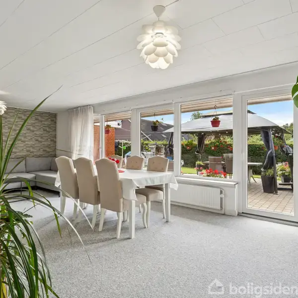 En spisebord med seks stole står foran store vinduer, der viser en terrasse med blomster og en have. Rummet er lyst og moderne indrettet med planter og lyse møbler.