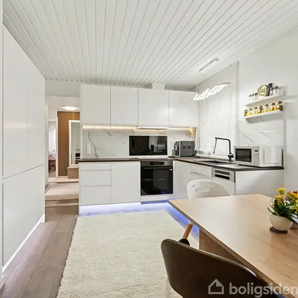 Et køkken med hvide skabe og apparater, sort kogeplade og ovn, i et lyst rum med træloft. Et træbord med blomsterdekoration står i forgrunden ved siden af et vindue.