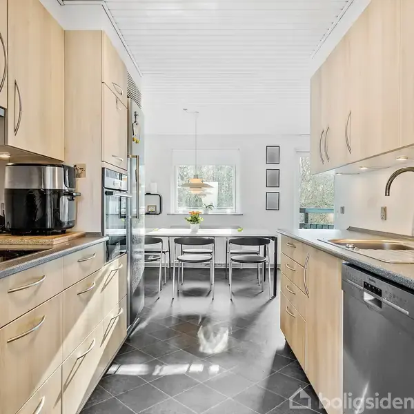 Et moderne køkken med lyse træskabe er udstyret med en kaffemaskine og opvaskemaskine. I baggrunden ses en spiseplads med et bord og fire stole nær et vindue.