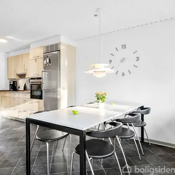 Moderne køkken med sort-hvid spisebord og stole. En plante og pendellampe pryder bordet. Baggrunden viser trægulve, skabe og et stort køleskab. Vægur viser klokken 10:10.