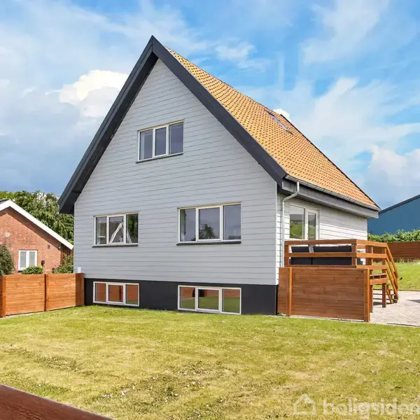 Hus står stille i en grøn have omkranset af et hegn. Det har en lysegrå facade og orange tag, med store vinduer og terrasse. Ingen tekst ses.