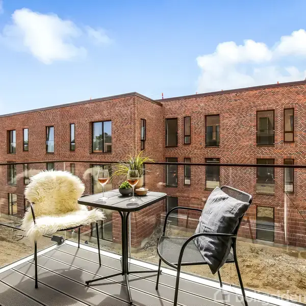 Et altanbord med to stole står stille på en moderne altan med glasgelænder, omgivet af murstensbygningens rolige, byggede kvarter. En vinflaske og glas på bordet tilføjer hygge.