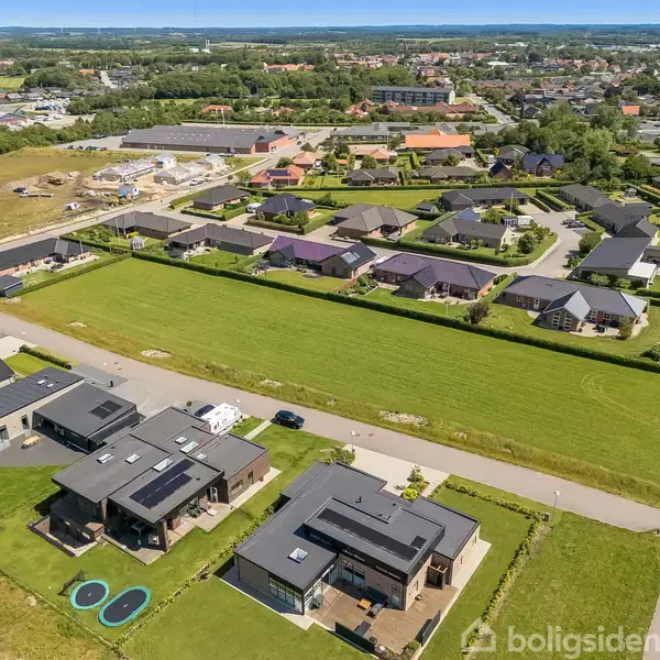 Husene med sorte tage ligger ved siden af en bred græsfelt i forstadsområde, omgivet af byggemodnede områder og flere boligkvarterer.