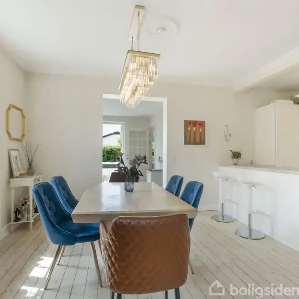 Et moderne køkken med et spisebord omgivet af blå og brun stole. En lysekrone hænger over bordet. Rummet har lyse vægge og gulve, med et stort vindue på venstre side.