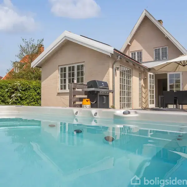 Udendørs jacuzzi med klart vand står foran en lys murstensvilla. I baggrunden ses en terrasse med grill og havemøbler under en parasol.