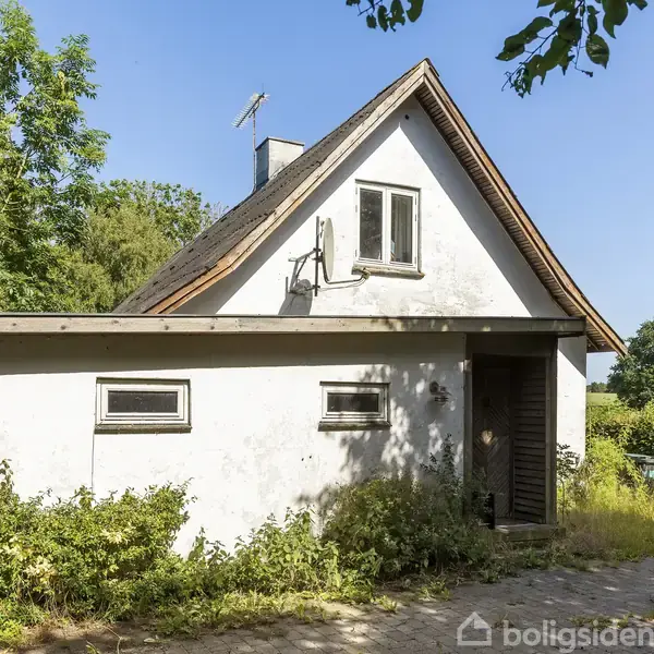 Et hvidt hus med skråt tag står omgivet af grønne træer og buske. Solen skinner på muren, og der er en skyggefuld indgang med dør.