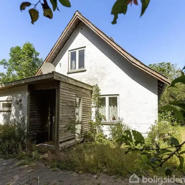 Et hvidt, to-etagers hus med skråt tag står omgivet af frodig vegetation. Huset har en træbeklædt indgang og flere vinduer. Omgivende træer og buske skaber en tæt natur omkring ejendommen.