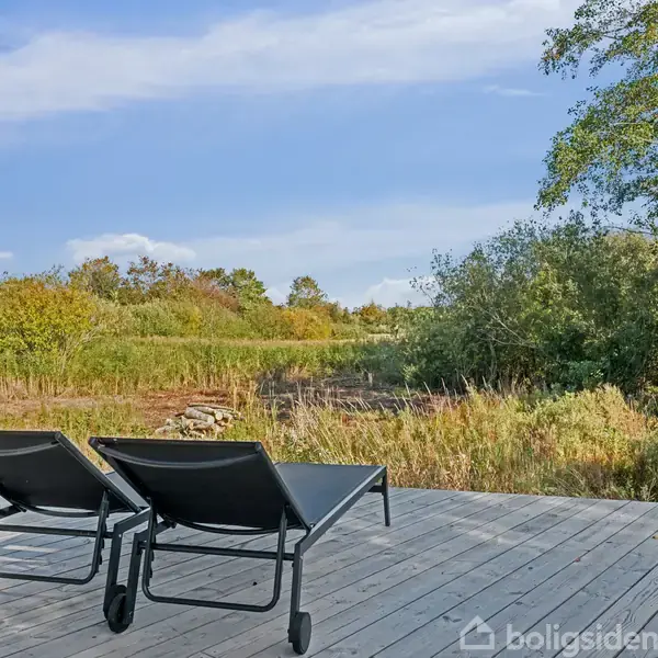 To liggestole står på en træterrasse. Foran dem er der en frodig, grøn natur med træer og buske under en blå himmel.