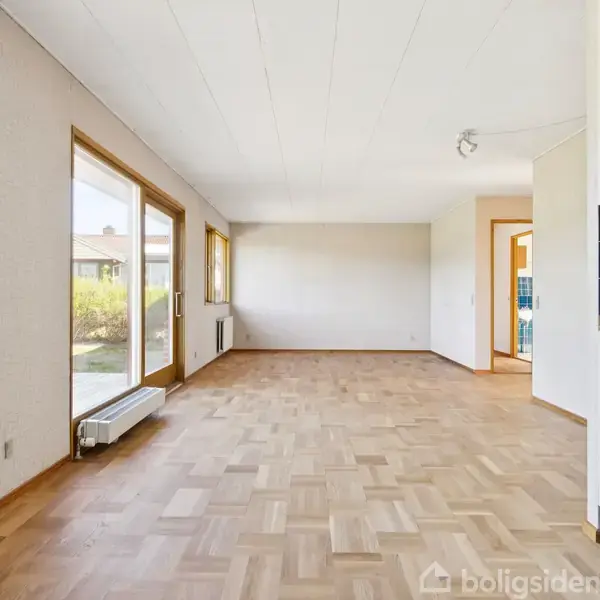 Stue med trægulv og hvidt loft. Der er store vinduer og en glasdør mod venstre, der giver udsigt til en have. Til højre ses et åbent køkkenområde med gule skabe.