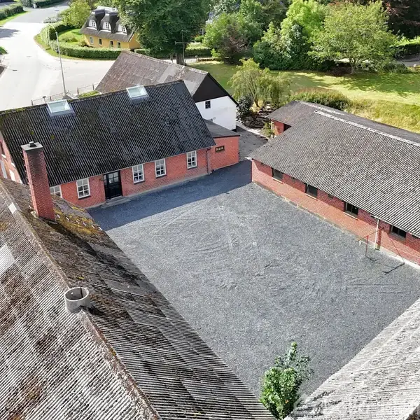 Luftfoto af en gård med røde murstensbygninger, der omkranser en grusbelagt gårdsplads. Omgivet af grønne områder med træer og en vej i nærheden.