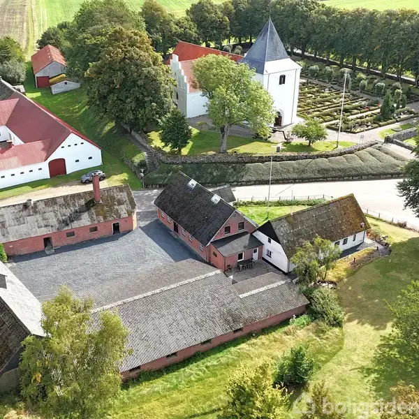 Gårde med røde tage står i en række ved siden af en hvid kirke med et rundt tårn, omgivet af træer og marker.