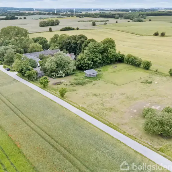 Landejendom omgivet af træer ligger ved lang smal vej, omgivet af marker med store åbne vidder i baggrunden og spredte vindmøller i horisonten under en overskyet himmel.
