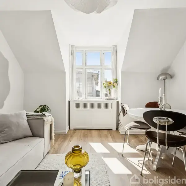 En lys stue med en moderne sofa til venstre, et stilfuldt spisebord til højre, og et stort vindue i midten, der tillader naturligt lys at strømme ind.