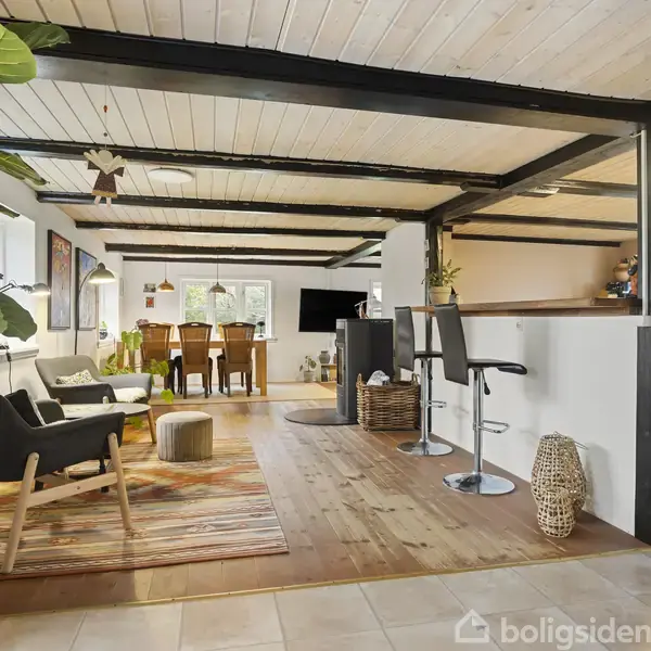 Stuen har træloft og bjælker, med loungeområde bestående af stole og et tæppe. Barstole står ved en disk. Der er et spisebord med stole i baggrunden.