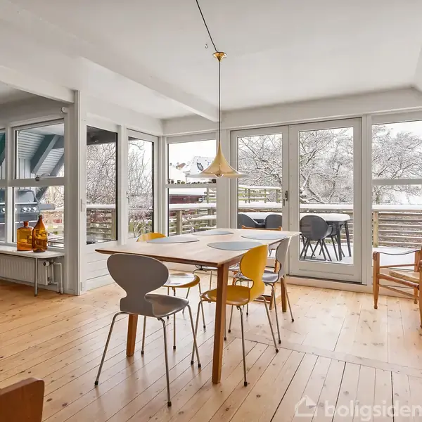 Et lyst spiseområde med et træbord omgivet af forskellige stole. Store vinduer viser en terrasse og træer udenfor. En lampe hænger over bordet, og rummet har trægulve.