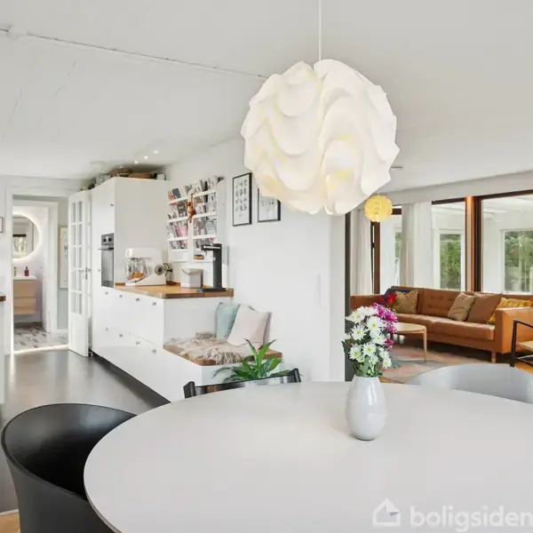 Rundt bord med vase og blomster står i moderne køkken-alrum. Store vinduer i baggrunden, lædersofa og gyngestol ses i stueområdet. Hylder og skabe er synlige i køkkenet.