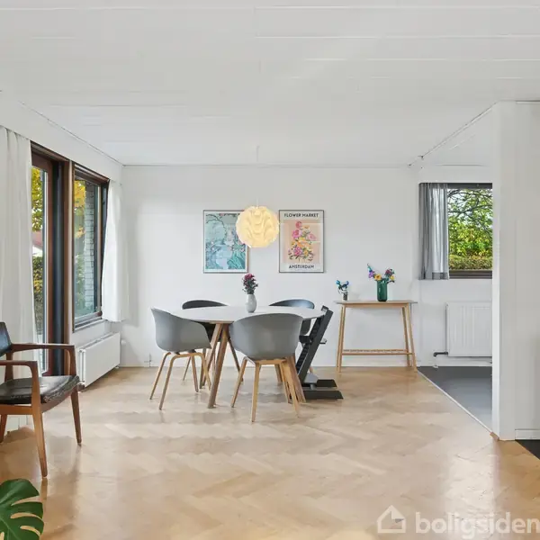 En moderne stue med et rundt bord omgivet af fire stole, ses i en lys indretning med trægulv. En pejs er tændt i hjørnet. Store vinduer slipper naturligt lys ind.