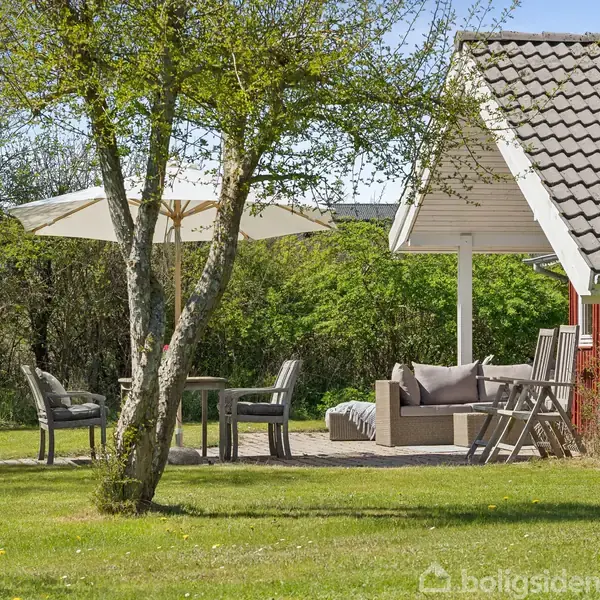 Et udendørs opholdsområde viser et træ, borde og stole under en parasol ved siden af et rødt hus. Haven er grøn og omkranset af buske og træer.