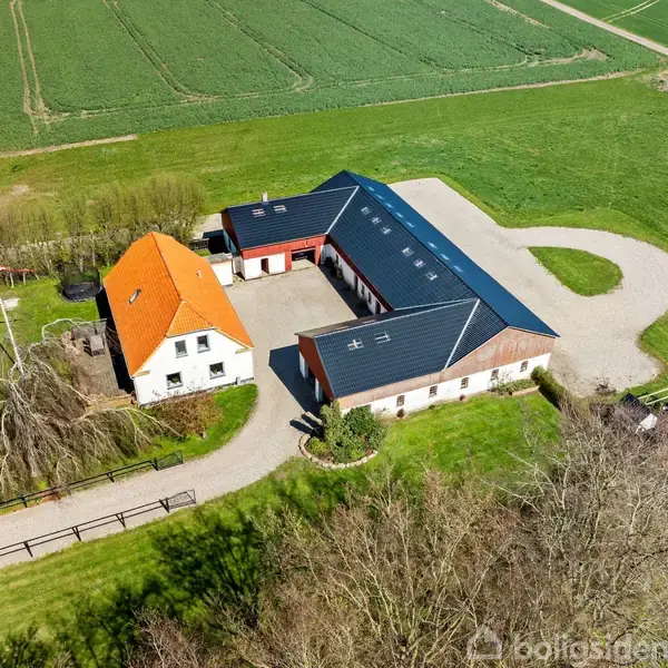 Gårdkompleks med et hvidt hus med orange tag placeret ved en landevej. En stor udbygning med sort tag omkranser en gårdsplads, omgivet af grønne marker.