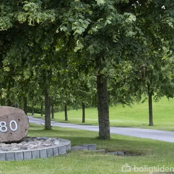 Sten med tallet "80" står på en rund platform, omgivet af grønne træer. En sti fører væk gennem parken.