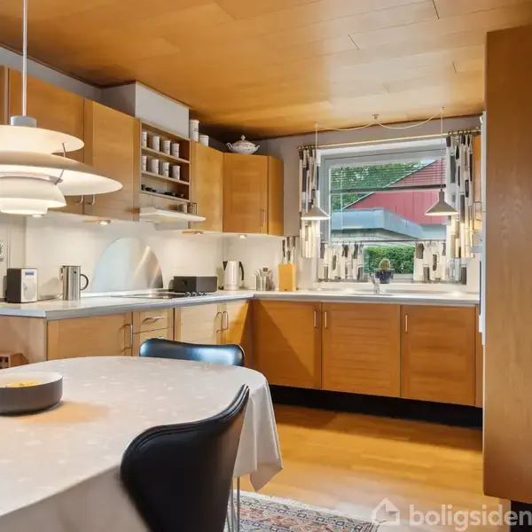 Et køkken med træskabe og hvide overflader har en spiseplads med et bord med dug og sorte stole. En pandehængt lampe hænger over bordet, og vinduet giver udsigt til en have.