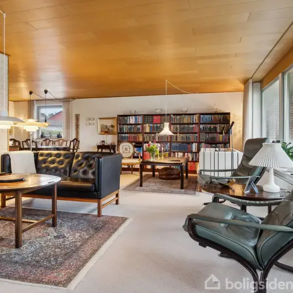 En stue med sorte lænestole og sofaer omkring et sofabord, belyst af moderne lamper. I baggrunden ses bogreoler fyldt med bøger, og store vinduer slipper lys ind.